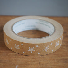 Load image into Gallery viewer, A roll of brown kraft paper tape printed with a pattern of white stars, resting on a wooden tabletop against a neutral gray background.