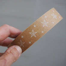 Load image into Gallery viewer, A hand holding a roll of brown kraft paper tape with white outlined star patterns, set against a plain gray background.