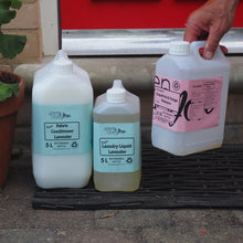Load image into Gallery viewer, Three large bottles of laundry detergent on a step with a red door in the background.