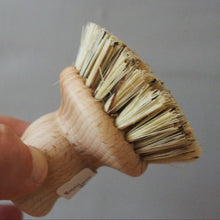 Load image into Gallery viewer, A hand holding a small round brush with natural wood bristles and a light-colored wooden handle against a plain gray background.