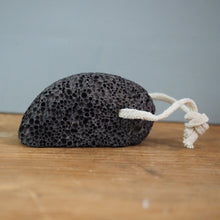 Load image into Gallery viewer, A black pumice stone with a white drawstring, resting on a wooden surface against a gray background.