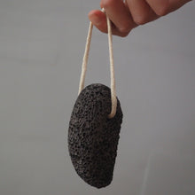 Load image into Gallery viewer, A dark gray porous lava stone bath sponge hanging from a light beige cotton cord held by a hand, set against a plain light gray background.