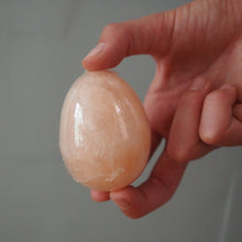 Load image into Gallery viewer, A hand holding a smooth, polished peach-colored himalayan salt egg against a plain gray background.