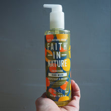 Load image into Gallery viewer, A hand holding a bottle of Faith In Nature Energising Hand Wash against a dark gray textured wall. The bottle is clear, showing a yellow liquid inside, with a white pump dispenser and a colorful label featuring orange, yellow, and blue graphics.