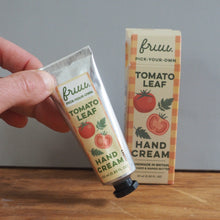 Load image into Gallery viewer, A hand holds a tube of Bruu Tomato Leaf Hand Cream next to its box on a wooden table against a gray wall.