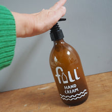 Load image into Gallery viewer, An amber glass hand cream bottle with a black pump dispenser, held by a person's hand wearing a green sweater. The bottle features a white printed logo and text reading 'fall HAND CREAM'.