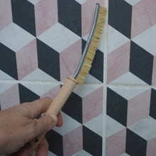 Load image into Gallery viewer, A hand holding a curved cleaning brush with light wood bristles and a silver metal edge, positioned against a geometric wall pattern of black, white, and pink tiles.