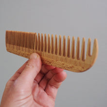 Load image into Gallery viewer, Hand holding a wooden comb against a plain background
