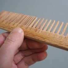 Load image into Gallery viewer, Hand holding a wooden comb against a gray background
