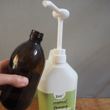 Load image into Gallery viewer, A hand holds a dark amber glass bottle above a white pump bottle of Bio Comfort Hand Cleansing Wash on a wooden surface.
