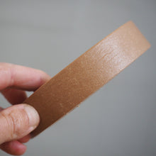 Load image into Gallery viewer, A hand holding a roll of light brown kraft paper tape against a plain gray background.
