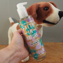 Load image into Gallery viewer, A hand holds a bottle of Faith in Nature dog shampoo in front of a plush beagle toy on a wooden floor against a gray wall.
