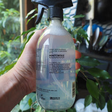 Load image into Gallery viewer, A hand holding a clear plastic spray bottle of Monstera Slickie cleaning spray, with a blurred indoor background featuring green plants and kitchen items.
