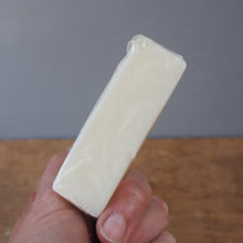 Load image into Gallery viewer, A hand holding a small rectangular cream-colored product wrapped in clear plastic, with a wooden surface on the left and a gray background on the right.
