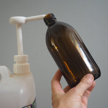 Load image into Gallery viewer, Person using a white pump to transfer liquid from a large container into a brown bottle against a gray background.
