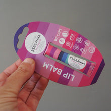 Load image into Gallery viewer, A hand holding a Benéanna lip balm tube in its original pink and purple cardboard hanger packaging against a plain gray background.
