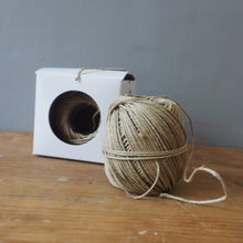 Load image into Gallery viewer, A ball of natural jute twine next to its white cardboard spool packaging, resting on a rustic wooden table against a plain gray wall.
