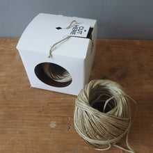 Load image into Gallery viewer, A white cardboard box with a circular cutout containing natural jute twine, next to a larger loose coil of the same twine on a wooden surface against a gray wall.
