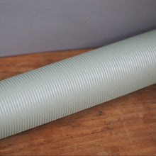 Load image into Gallery viewer, A light sage green corrugated flexible pipe or hose resting diagonally on a wooden surface against a gray background.
