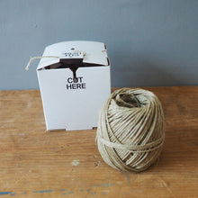 Load image into Gallery viewer, A ball of light tan jute twine next to its white cardboard packaging box on a rustic wooden table against a gray wall.
