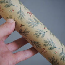 Load image into Gallery viewer, A hand holding a roll of kraft paper wrapping paper with a festive pattern of green pine branches, small red berries, and red bows against a solid gray background.
