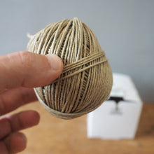 Load image into Gallery viewer, A hand holding a small ball of natural jute twine, with a white box blurred in the background on a wooden surface.
