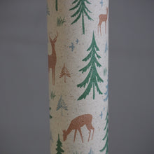Load image into Gallery viewer, A roll of wallpaper featuring a woodland pattern with brown deer, green pine trees, and small blue birds on a speckled cream background, shown against a solid gray backdrop.
