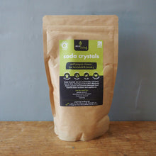 Load image into Gallery viewer, A stand-up kraft paper pouch of 'soda crystals' by the brand 'The Sodbird', standing on a wooden surface against a gray wall.
