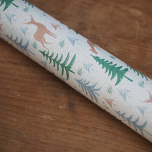 Load image into Gallery viewer, A close-up of a cylindrical drinking straw with a festive woodland print, featuring green pine trees, blue evergreens, and brown deer on a cream-colored background, resting on a wooden surface.
