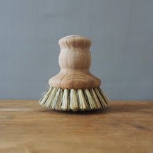 Load image into Gallery viewer, A small wooden cleaning brush with a light natural wood handle and natural straw-colored bristles, standing on a wooden tabletop against a plain gray wall.
