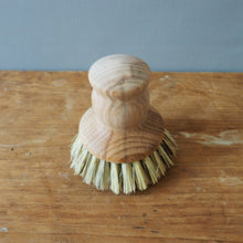 Load image into Gallery viewer, A small wooden cleaning brush with a light natural wood handle and pale straw-colored bristles, standing on a worn wooden tabletop against a plain gray wall.
