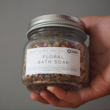 Load image into Gallery viewer, A hand holding a clear glass jar of Organic Living Floral Bath Soak with a white label and silver metal lid against a plain gray background.
