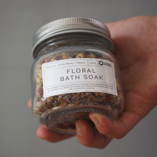Load image into Gallery viewer, A hand holding a clear glass jar of floral bath soak with a silver lid and a white label, set against a plain gray background.
