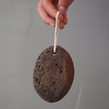 Load image into Gallery viewer, A hand holding a dark brown pumice stone bath sponge with a white hanging loop against a plain gray background.
