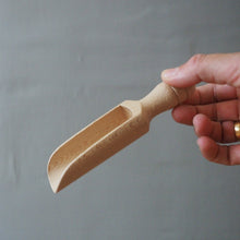 Load image into Gallery viewer, A hand holding a small, unfinished wooden scoop or garden tool against a gray wall and wooden tabletop background.
