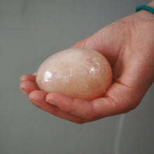 Load image into Gallery viewer, A hand holding a smooth, oval-shaped pale pink himalayan salt egg or crystal against a plain gray background.
