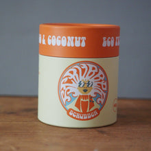 Load image into Gallery viewer, A cylindrical jar of Schabbz Coconut &amp; Clay Clay Mask, featuring an orange lid and a cream-colored body with a colorful tree logo, set on a wooden surface against a gray background.
