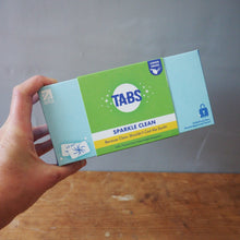 Load image into Gallery viewer, A hand holding a box of Tars Sparkle Clean Multi-Use Clean Powder against a split background of a wooden table on the left and a gray surface on the right.
