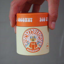 Load image into Gallery viewer, A hand holding a small jar of coconut egg scrub against a plain gray background. The jar has an orange lid with white text and a cream-colored body featuring a colorful circular logo.
