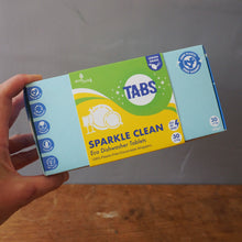 Load image into Gallery viewer, A hand holding a box of Sparkle Clean Eco Dishwasher Tablets by The Dishy Dishes brand, set against a plain gray wall and wooden floor background.
