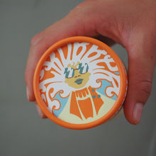 Load image into Gallery viewer, A hand holding a round container with a bright orange lid featuring a stylized sun and face illustration in orange, white, blue, and yellow against a plain gray background.
