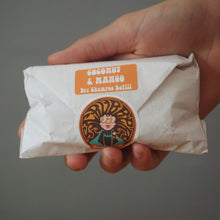 Load image into Gallery viewer, A hand holding a crumpled white paper-wrapped bar or stick product with an orange label reading 'COCONUT &amp; MANGO' and 'Sri Shanmuk Kellass' plus a circular illustrated logo, against a plain gray background.
