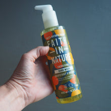 Load image into Gallery viewer, A hand holding a bottle of Faith In Nature Energizing Hand Wash against a dark gray background. The bottle contains a yellow liquid and features a white pump dispenser with a colorful label.

