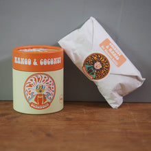 Load image into Gallery viewer, A container of Schumacher Mango &amp; Coconut sweets stands on a wooden surface against a gray wall, next to a white paper bag with a matching circular logo.
