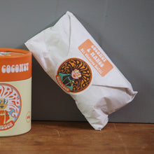 Load image into Gallery viewer, A cylindrical container of Scrubber Mango &amp; Coconut spread next to a white paper mailer envelope, both featuring the brand's circular logo, set on a wooden surface against a gray wall.
