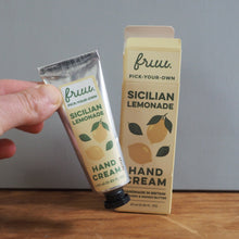 Load image into Gallery viewer, A hand holds a tube of Frui Sicilian Lemonade hand cream next to its packaging box on a wooden surface against a gray wall.
