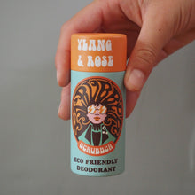 Load image into Gallery viewer, A hand holding a small cylindrical scrubber deodorant container against a plain gray background. The container has an orange cap with white text reading 'Ylang &amp; Rose' and a light blue body with a large illustrated label featuring orange and black details.
