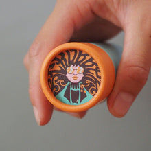 Load image into Gallery viewer, A hand holding the circular end of a cylindrical product, which features an orange rim and a printed illustration of a stylized face with dark hair on a teal background.
