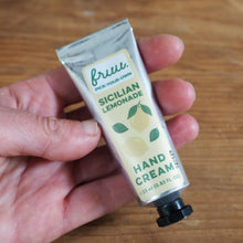 Load image into Gallery viewer, A hand holding a squeeze tube of Bruss Sicilian Lemonade Hand Cream against a wooden tabletop background.
