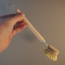 Load image into Gallery viewer, A hand holding a small cleaning brush with a light wood handle, metal wire hook at the end, and cream-colored bristles against a plain beige background.
